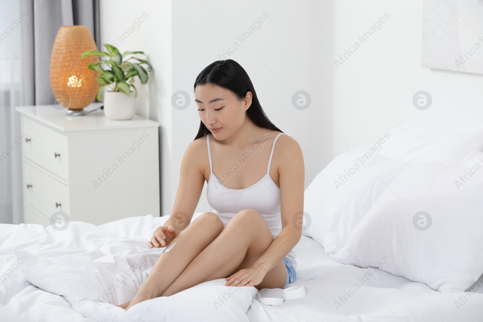 Photo of Beautiful young Asian woman applying body cream on leg in bedroom