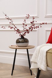 Photo of Hawthorn branches with red berries in vase on table near white wall indoors