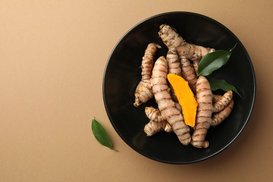 Photo of Bowl with raw turmeric roots and green leaves on light brown table, flat lay. Space for text