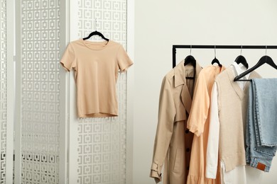 Photo of Beige t-shirt hanging on folding screen near rack with stylish women's clothes indoors