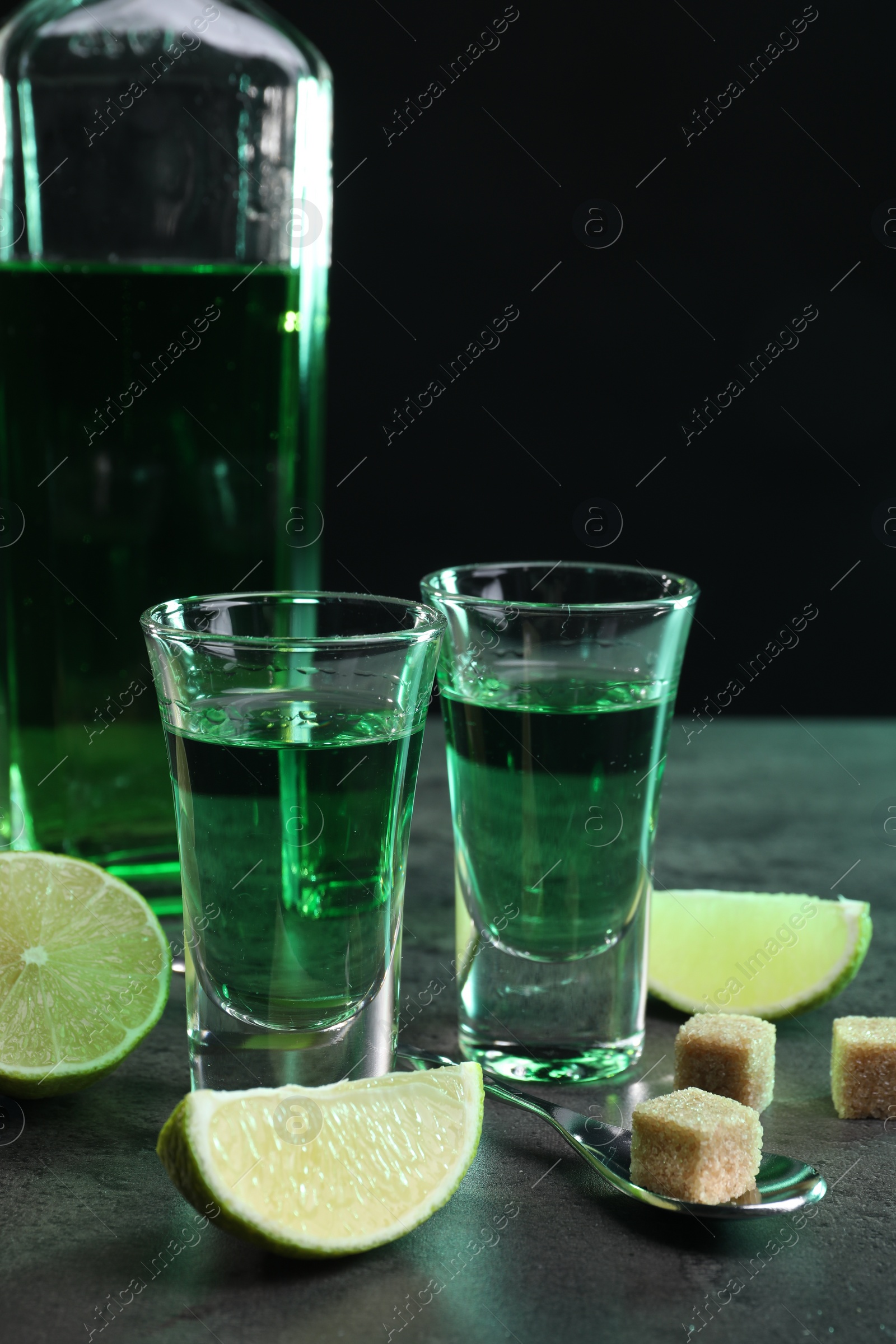 Photo of Absinthe in shot glasses, spoon, brown sugar cubes and lime on gray textured table, closeup. Alcoholic drink