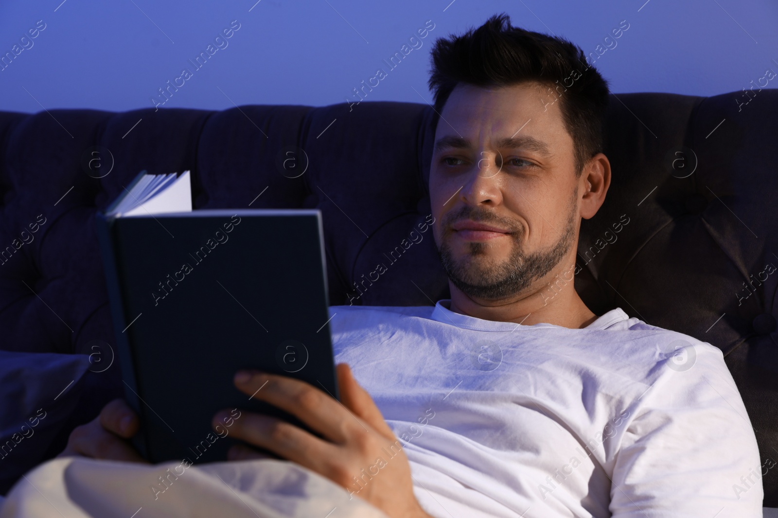 Photo of Handsome man reading book in dark room at night. Bedtime