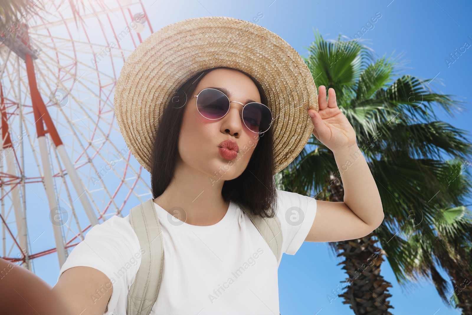 Image of Smiling young woman in sunglasses and straw hat taking selfie near observation wheel