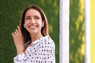 Young woman with wireless headphones listening to music near green grass wall. Space for text