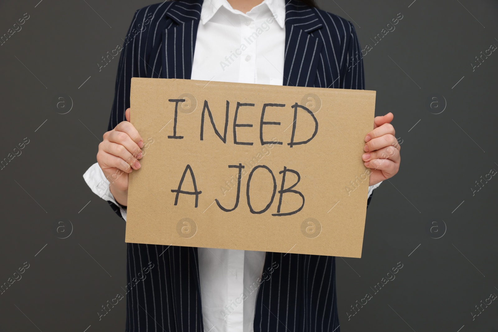 Photo of Young unemployed woman holding sign with phrase I Need A Job on black background, closeup