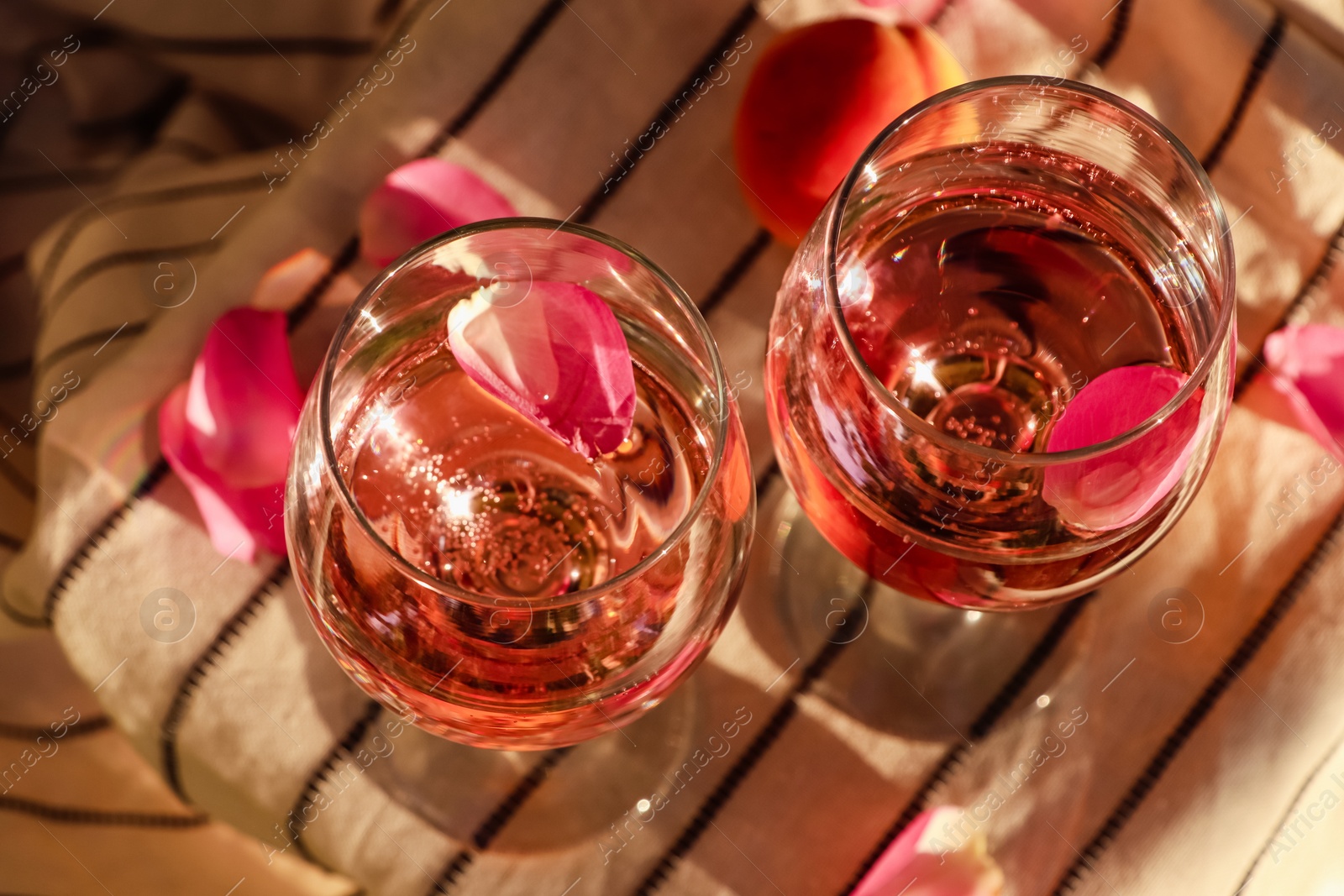 Photo of Glasses of delicious rose wine with petals and peaches on white picnic blanket outside, above view