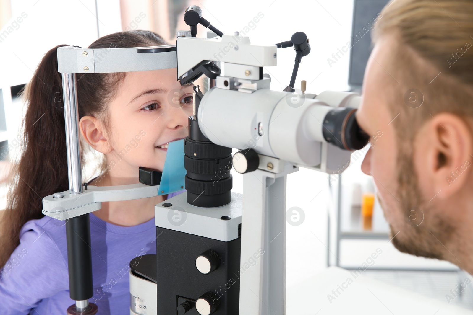 Photo of Children's doctor examining little girl with ophthalmic equipment in clinic