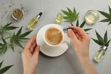 Photo of Top view of woman dripping THC tincture or CBD oil into coffee at light grey table, closeup