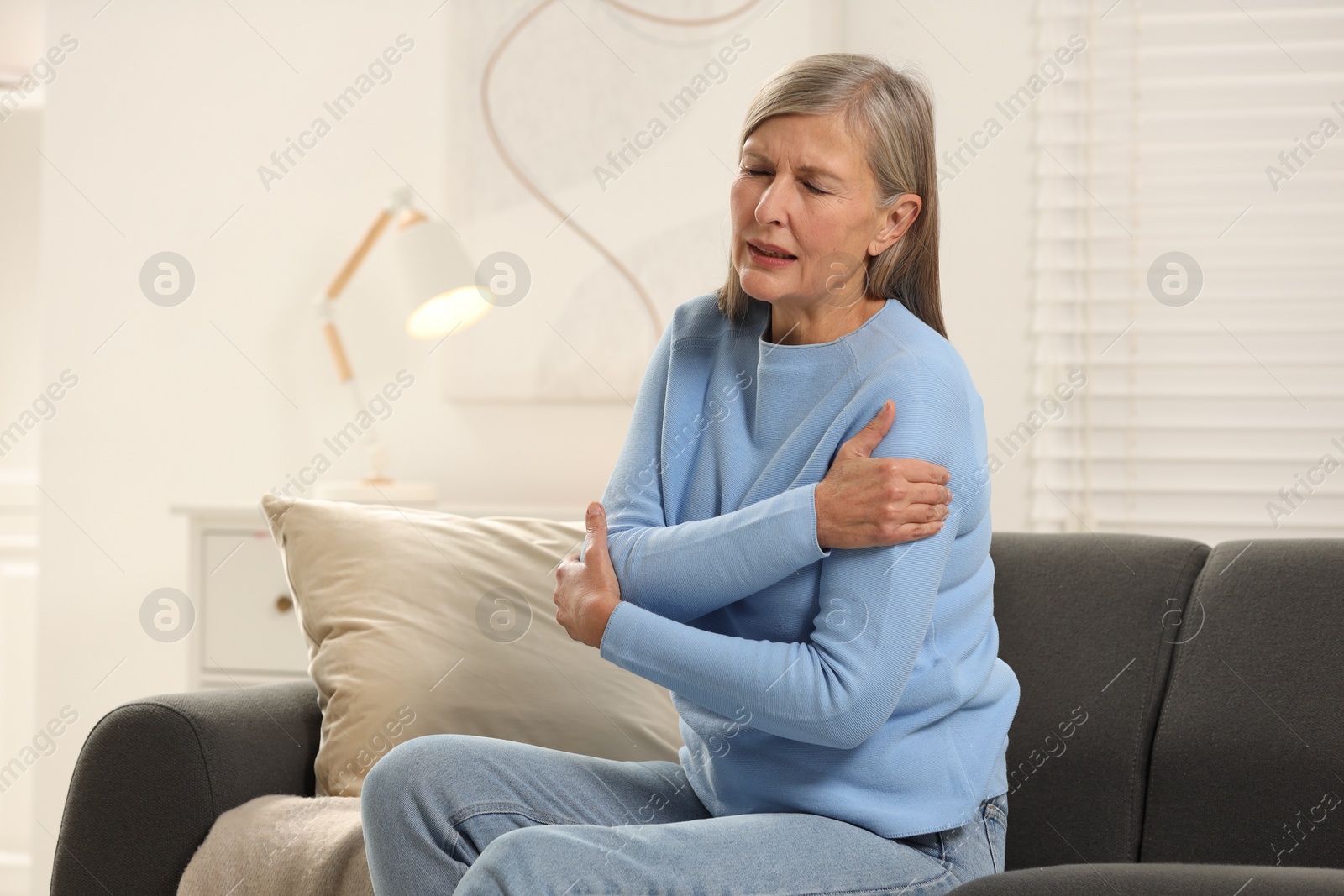 Photo of Mature woman suffering from pain in arm on sofa at home. Rheumatism symptom