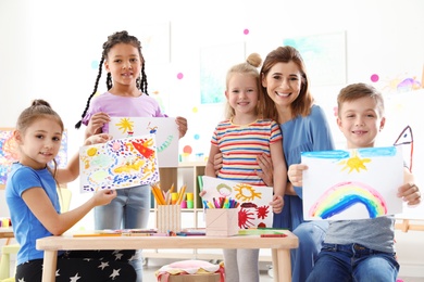 Photo of Cute little children with teacher showing their paintings at lesson indoors