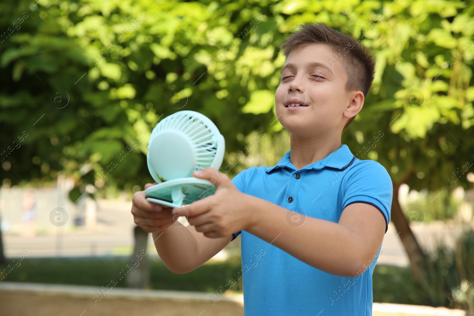 Photo of Little boy enjoying air flow from portable fan outdoors. Summer heat