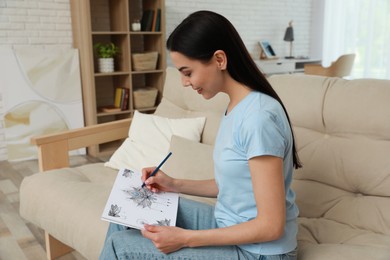 Photo of Young woman coloring antistress page on sofa in living room