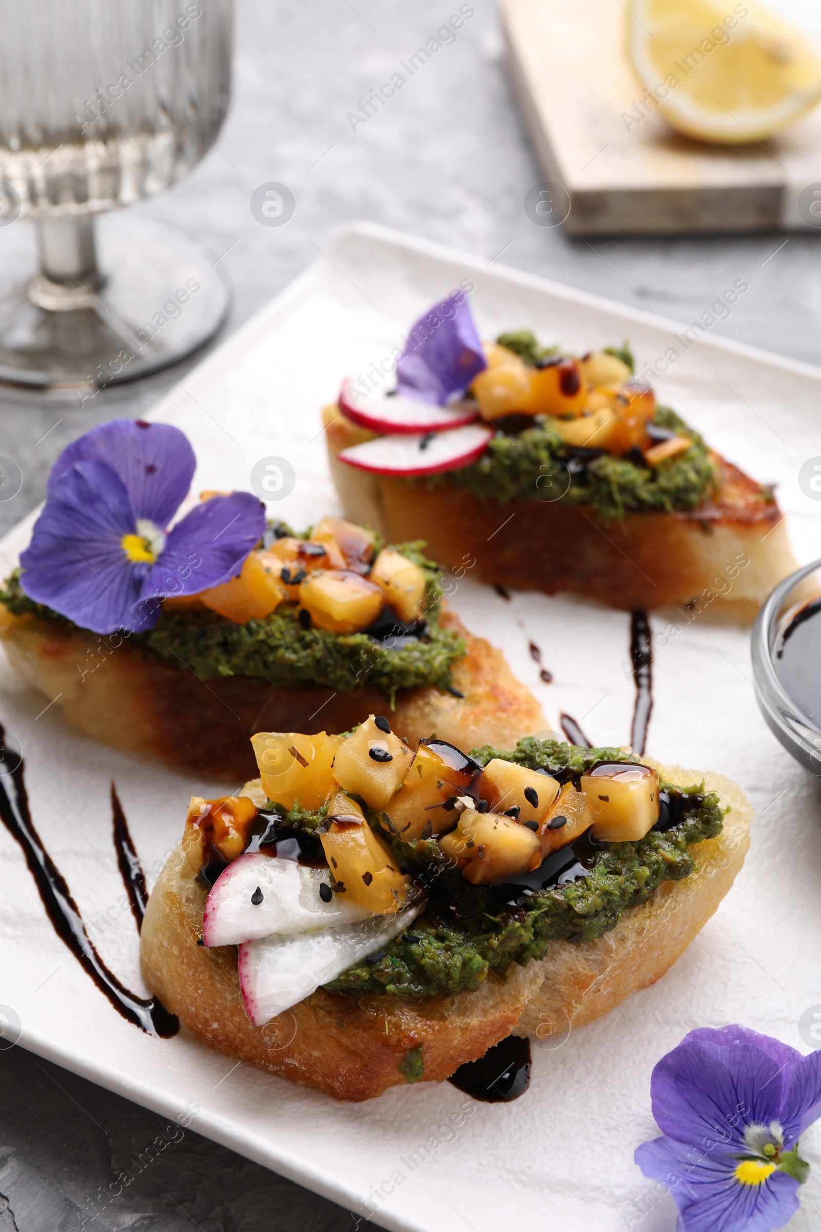 Photo of Delicious bruschettas with pesto sauce, tomatoes, balsamic vinegar and violet flowers on gray table, closeup
