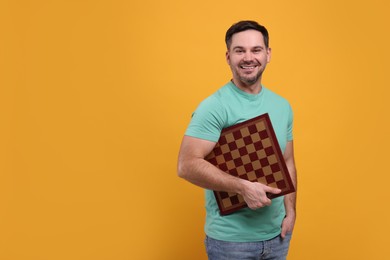 Smiling man holding chessboard on orange background, space for text