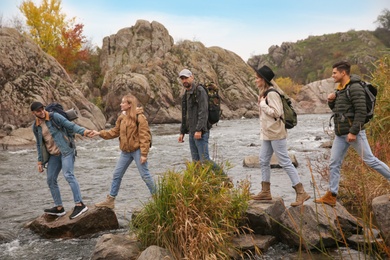 Group of friends with backpacks crossing mountain river on autumn day