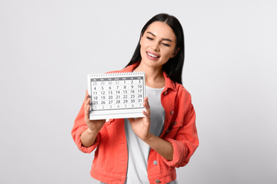 Young woman holding calendar with marked menstrual cycle days on light background