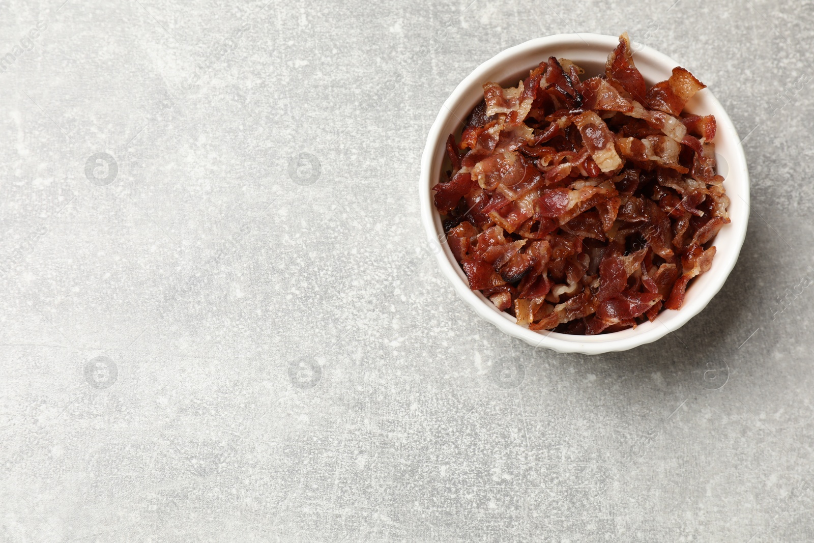 Photo of Pieces of tasty fried bacon in bowl on gray table, top view. Space for text