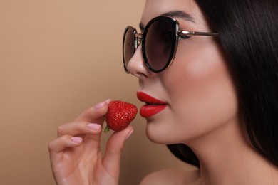 Photo of Attractive woman in fashionable sunglasses holding strawberry against beige background, closeup. Space for text