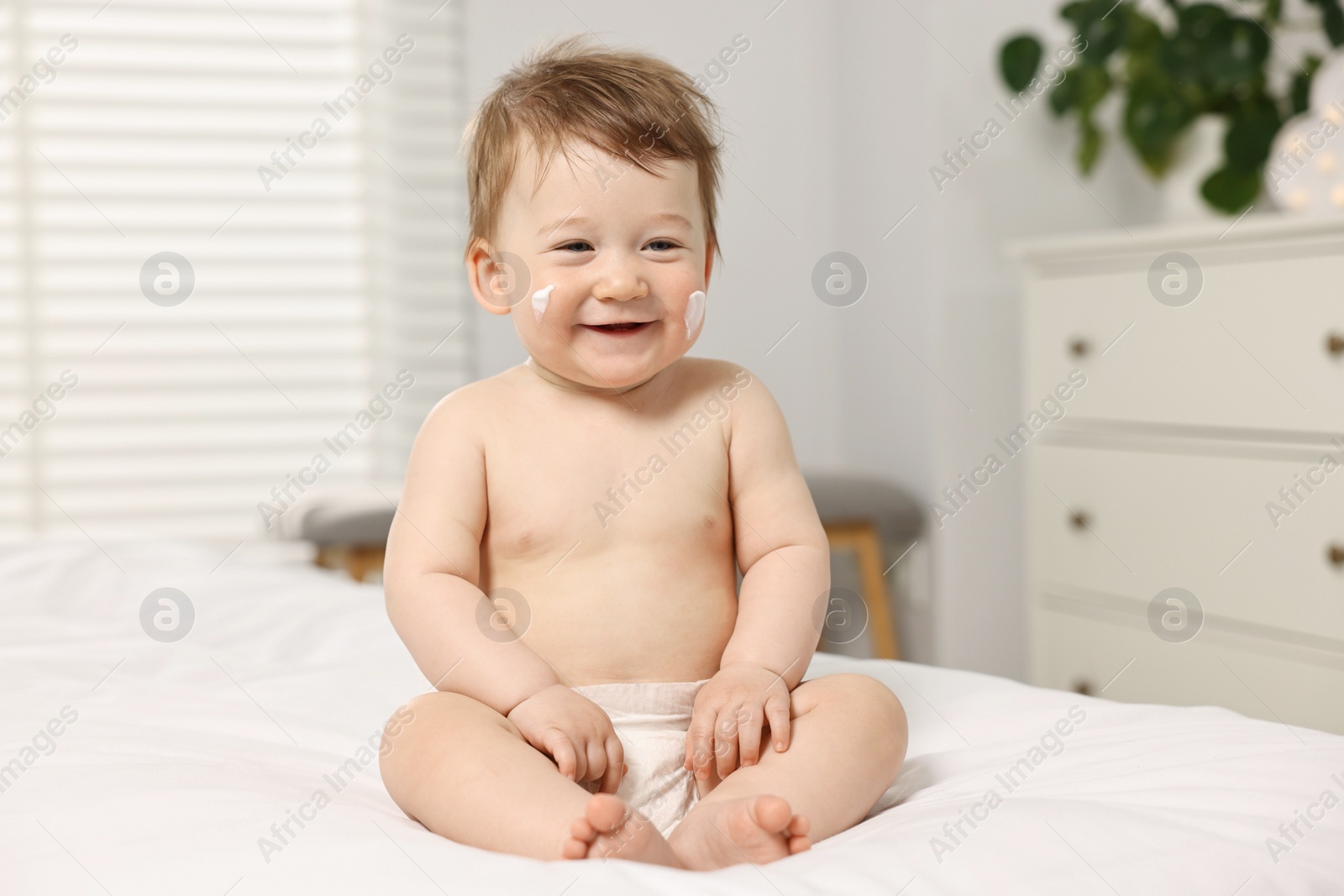 Photo of Cute little baby with moisturizing cream on face sitting on bed at home