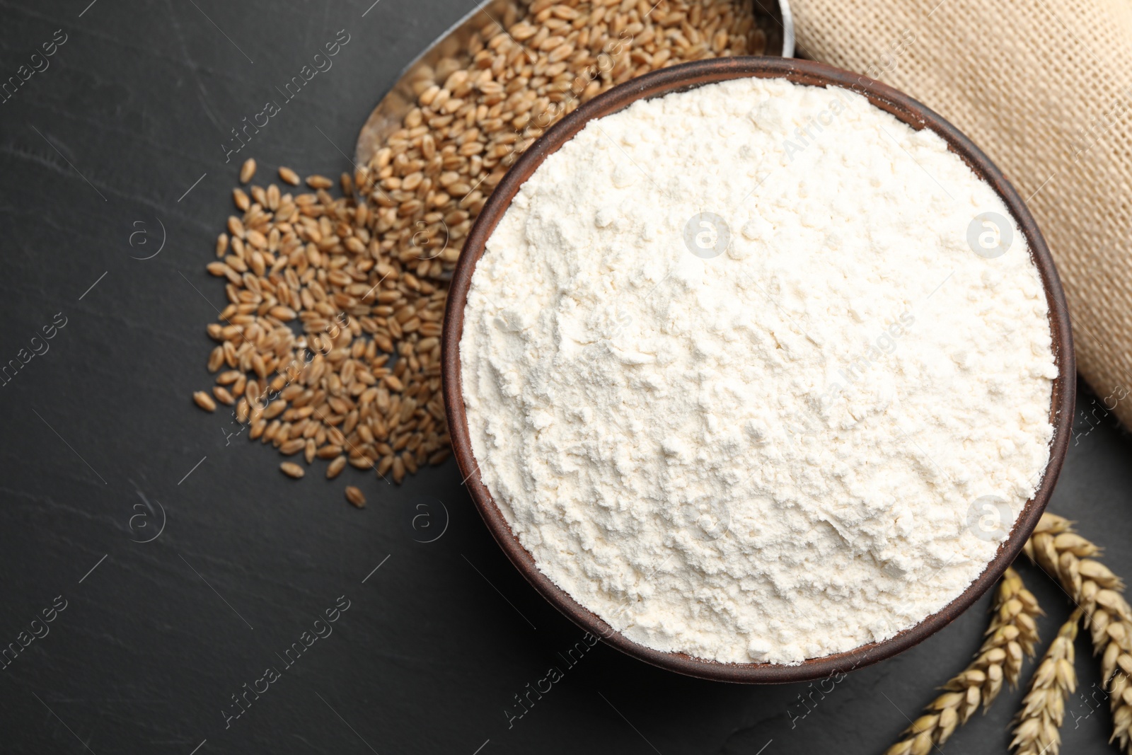 Photo of Wheat flour in bowl, spikes and grains on black table, flat lay. Space for text