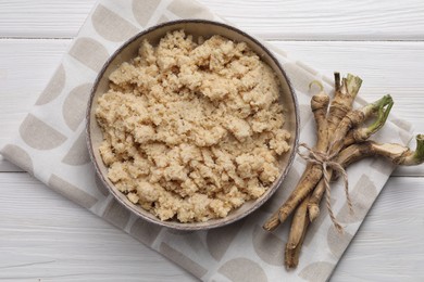Photo of Bowl of tasty prepared horseradish and roots on white wooden table, flat lay