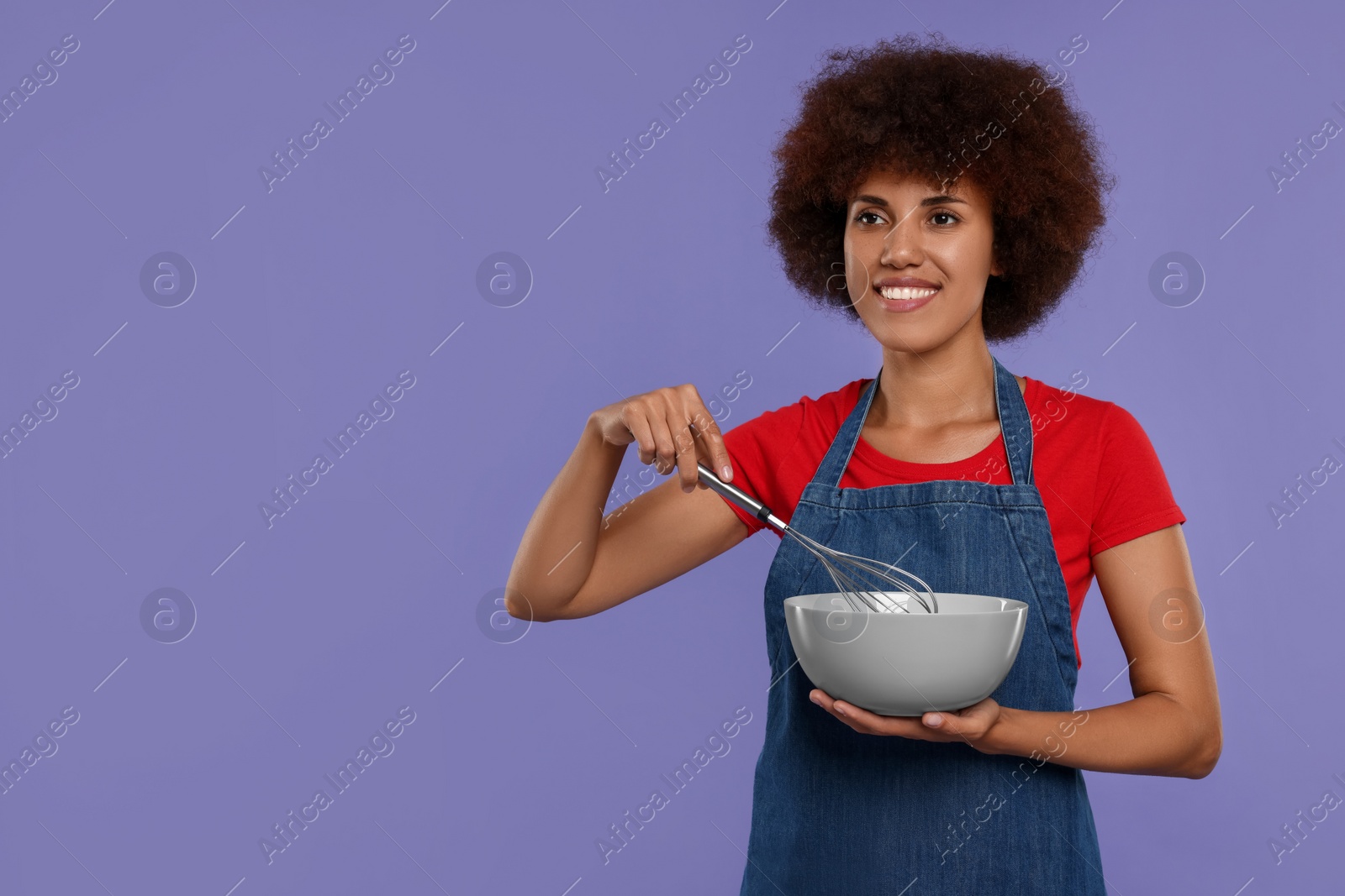 Photo of Happy young woman in apron with whisk and bowl on purple background. Space for text