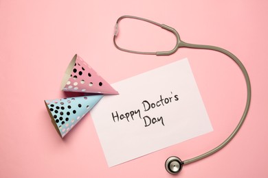 Photo of Card with phrase Happy Doctor's Day, stethoscope and party hats on pink background, flat lay