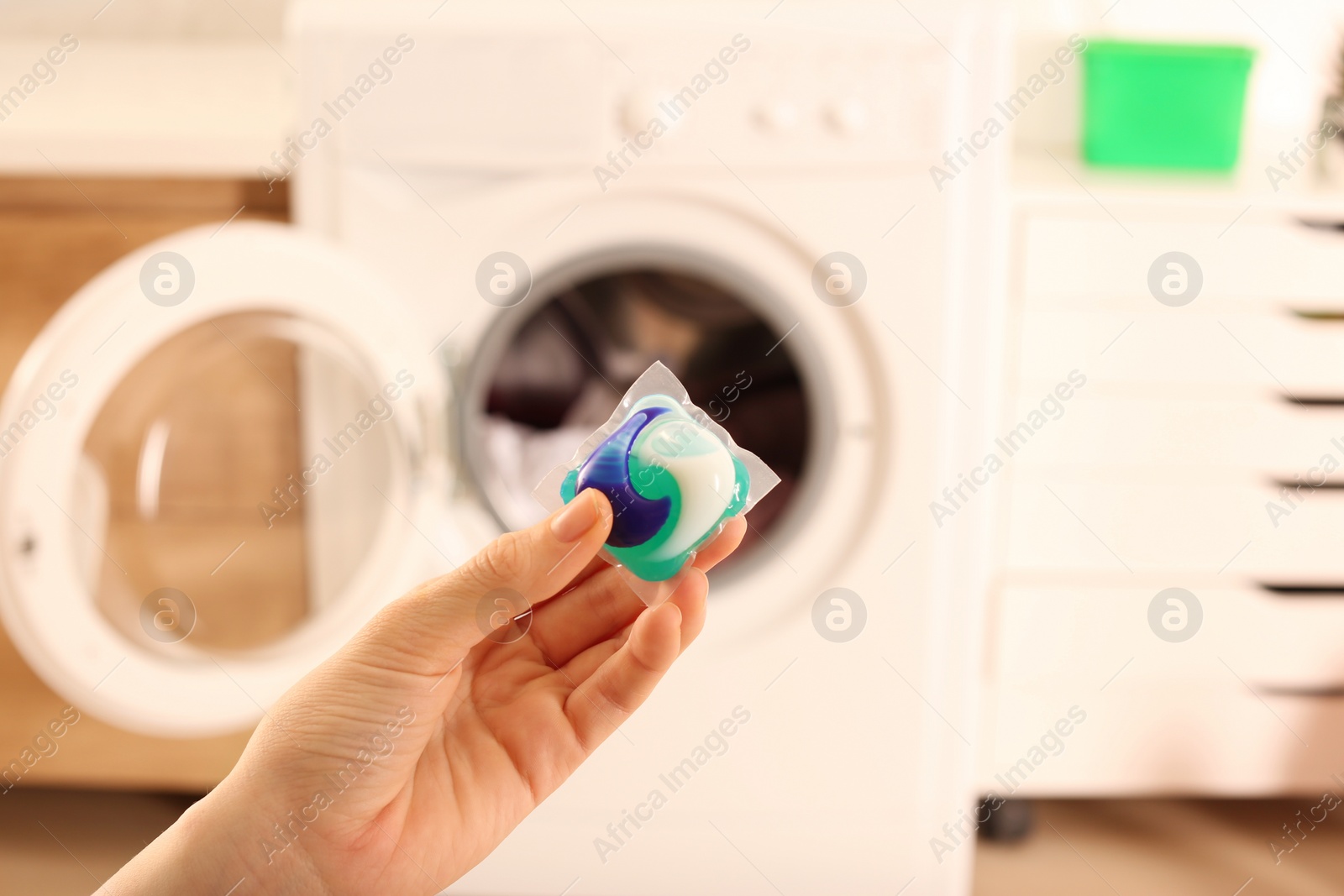 Photo of Woman holding laundry detergent capsule near washing machine indoors, closeup