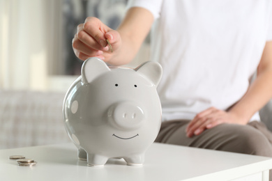 Photo of Woman putting money into piggy bank at white table indoors, closeup