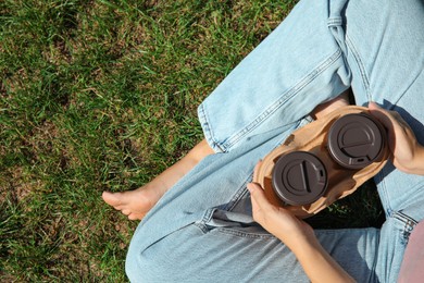 Photo of Woman holding takeaway cardboard coffee cups with plastic lids on green grass, top view. Space for text