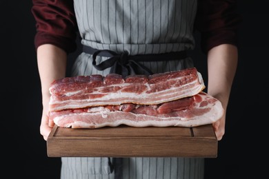 Photo of Woman holding wooden board with pieces of raw pork belly on black background, closeup