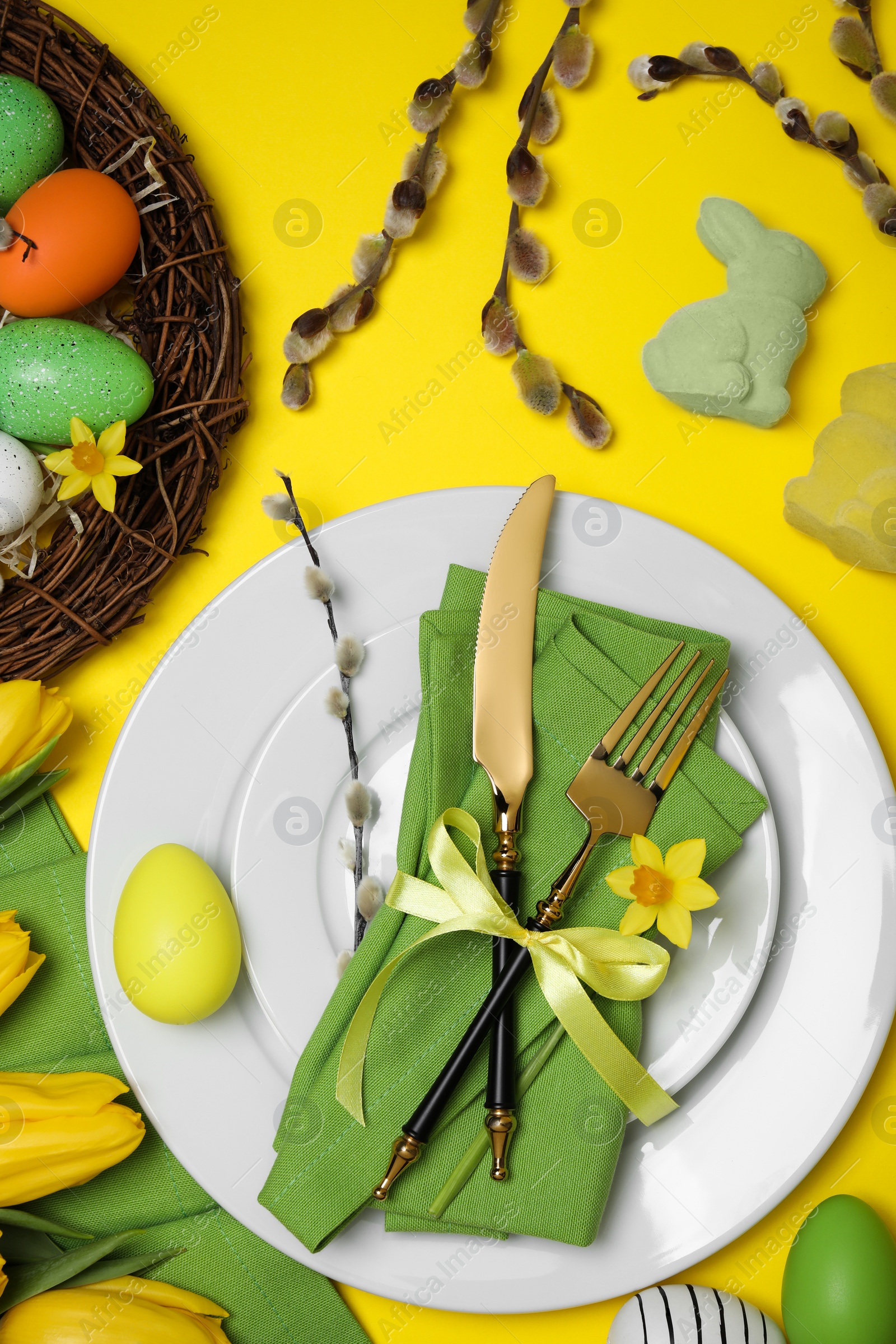 Photo of Festive table setting with painted eggs and tulips on yellow background, flat lay. Easter celebration