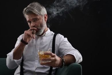Bearded man with glass of whiskey smoking cigar on sofa against black background. Space for text