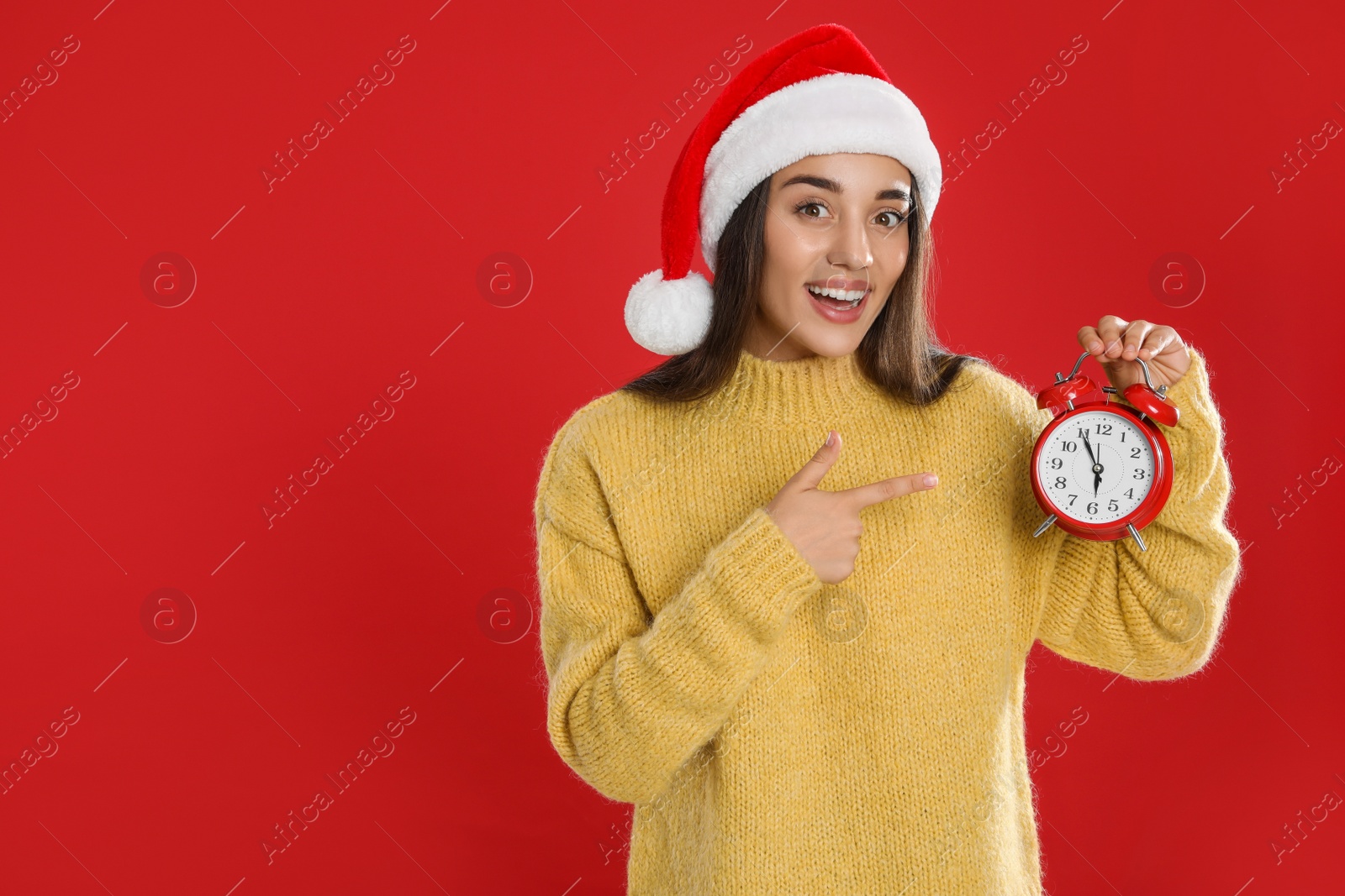 Photo of Woman in Santa hat with alarm clock on red background, space for text. New Year countdown