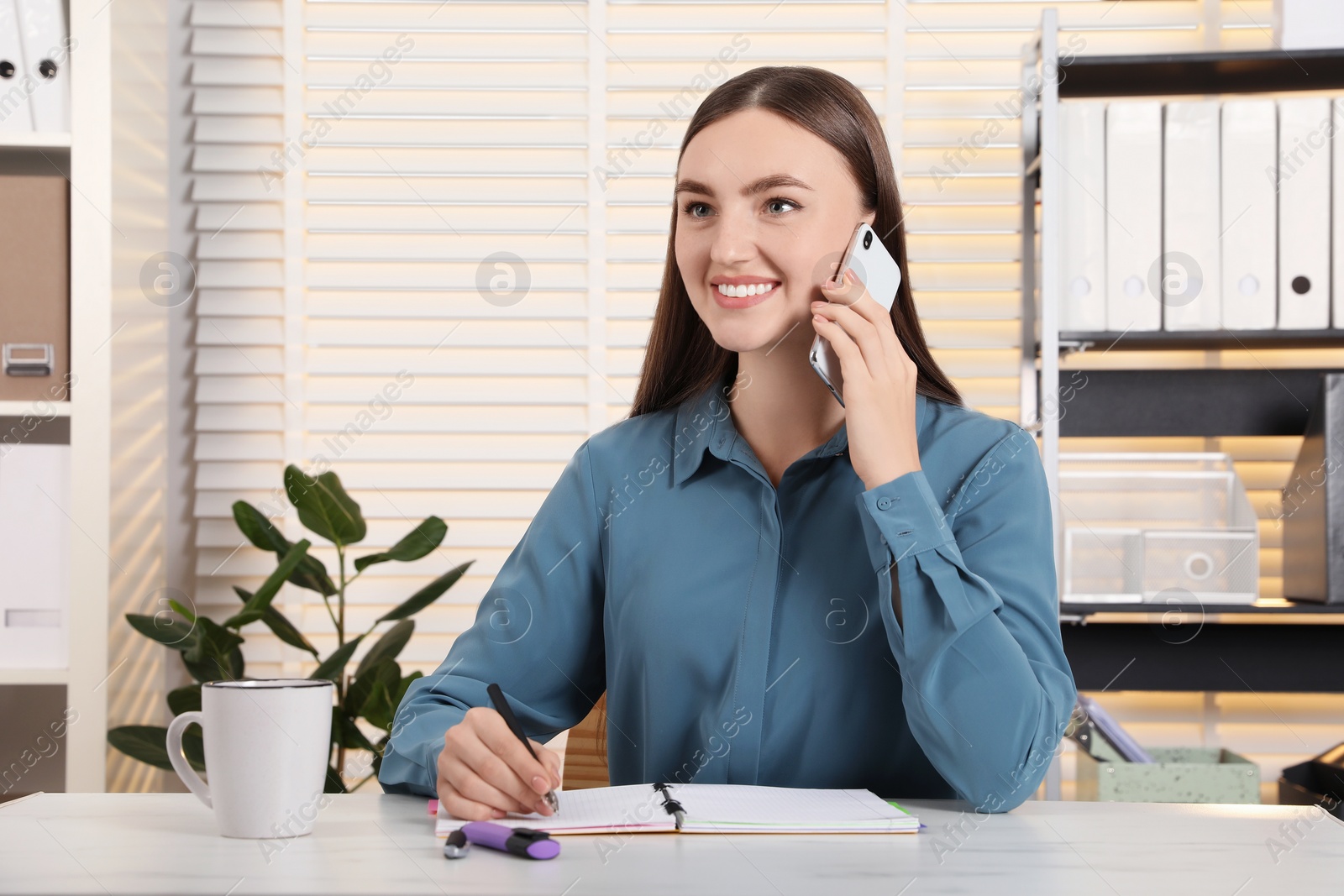 Photo of Happy woman taking notes while talking on smartphone at white marble table in office