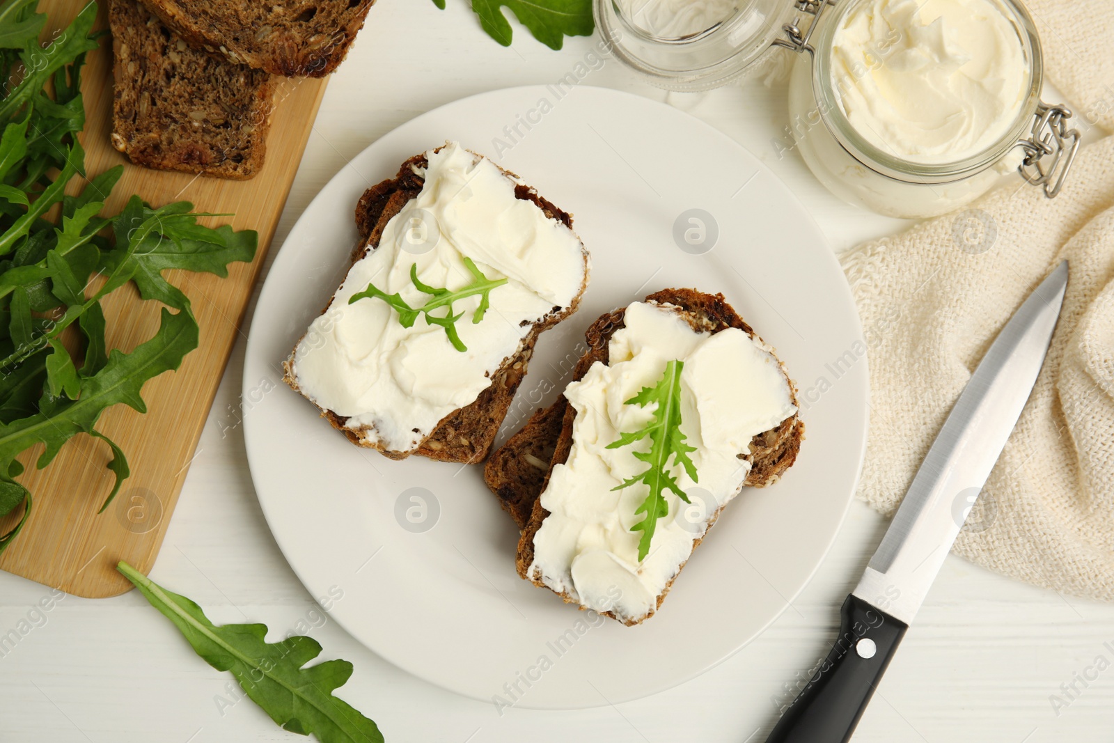 Photo of Bread with cream cheese and arugula on white wooden table, flat lay