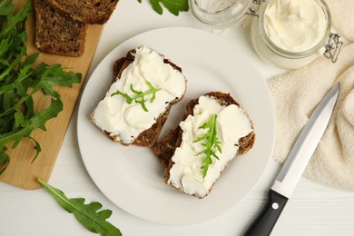 Bread with cream cheese and arugula on white wooden table, flat lay