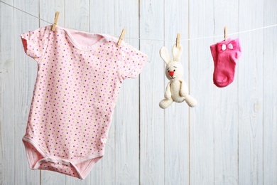 Photo of Bodysuit, pair of socks and toy bunny on laundry line against wooden background. Baby accessories