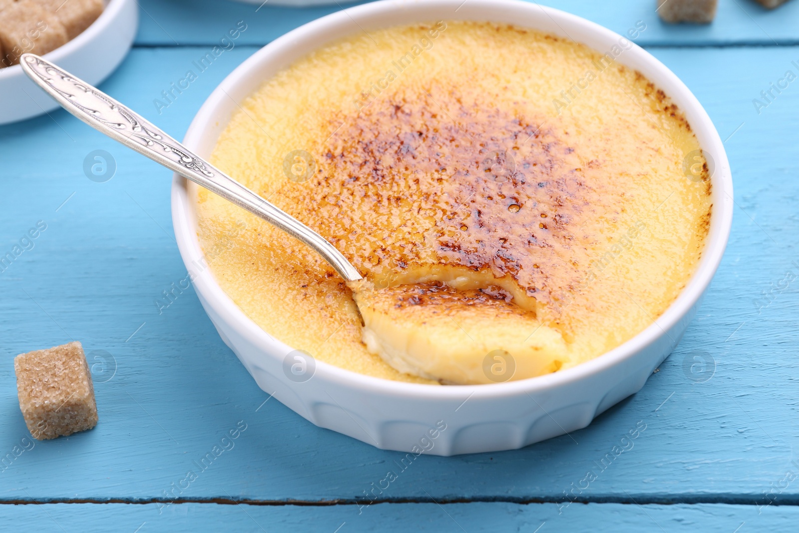 Photo of Delicious creme brulee in bowl, sugar cube and spoon on light blue wooden table, closeup