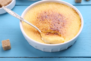 Photo of Delicious creme brulee in bowl, sugar cube and spoon on light blue wooden table, closeup