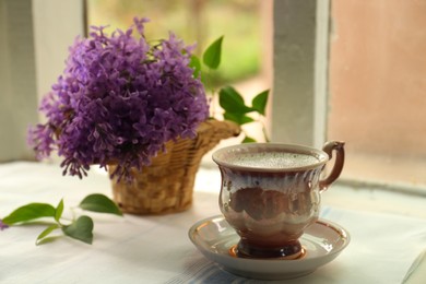 Beautiful lilac flowers in wicker basket and cup of hot coffee on window sill indoors