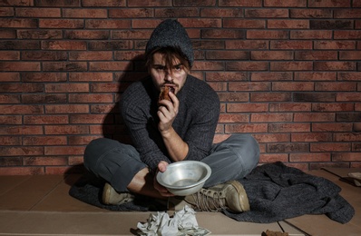 Poor young man with bread on floor near brick wall