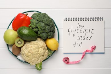 Notebook with words Low Glycemic Index Diet, measuring tape and healthy products on white wooden table, flat lay