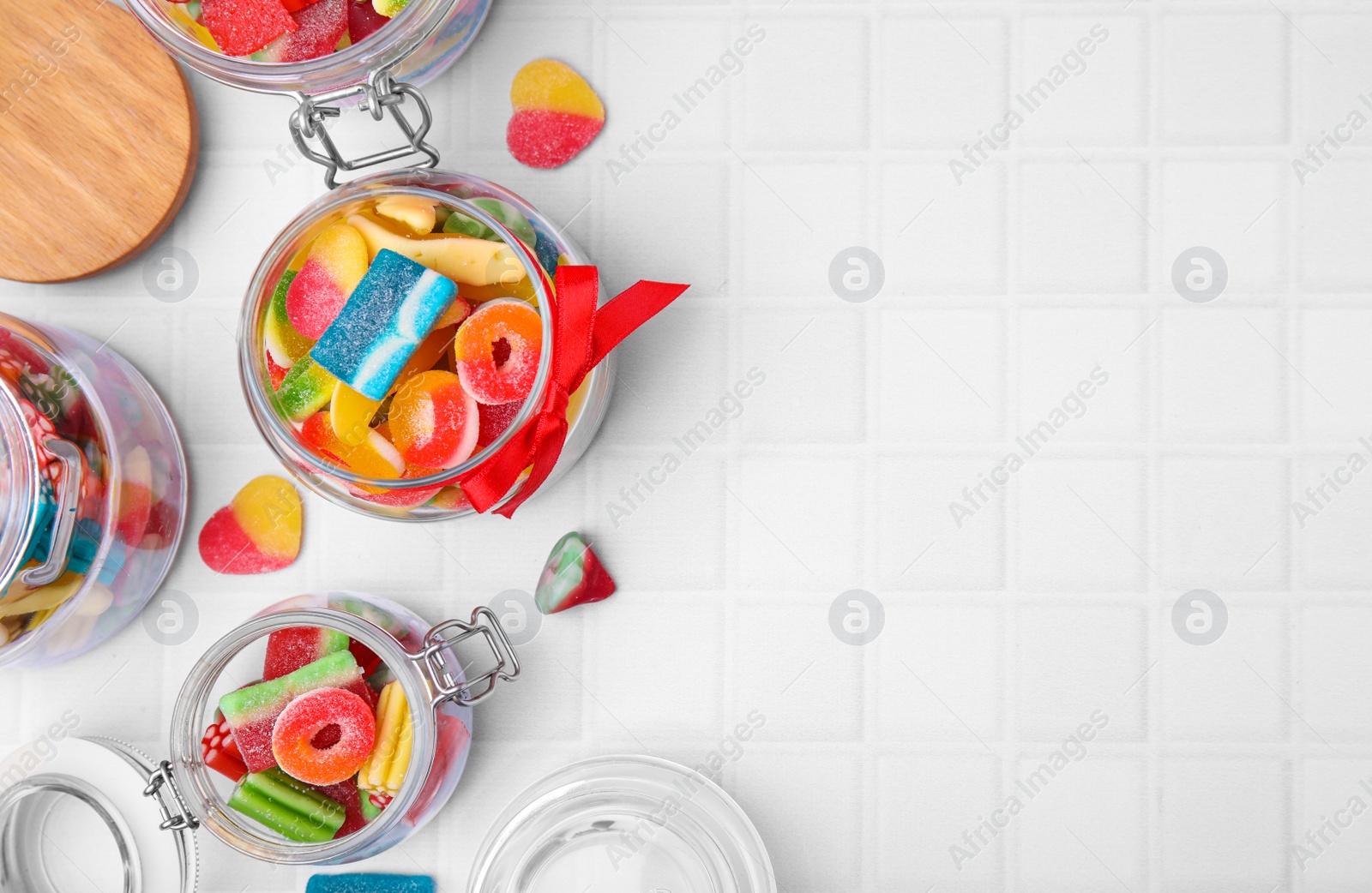 Photo of Glass jars with tasty colorful jelly candies on white tiled table, flat lay. Space for text