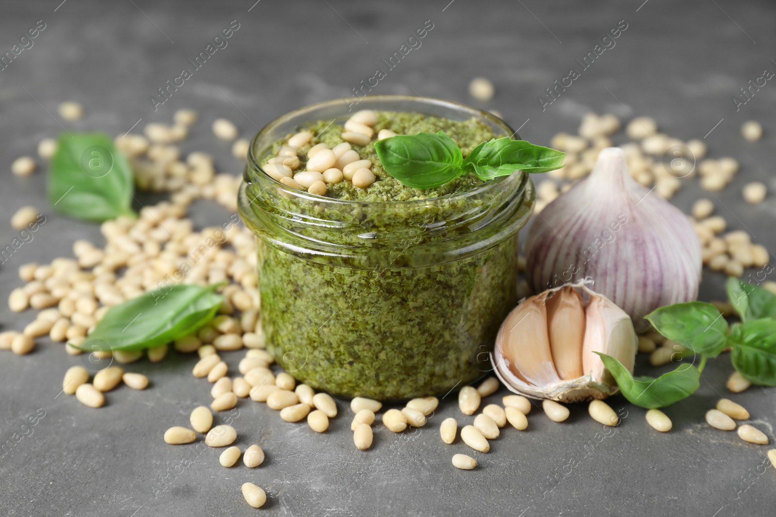 Photo of Jar of delicious pesto sauce and ingredients on grey table, closeup