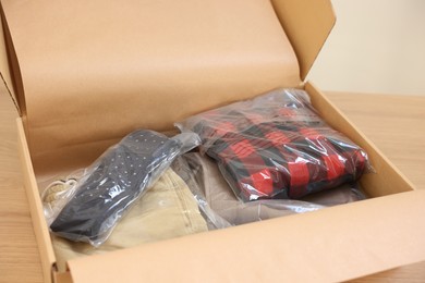 Photo of Open cardboard box with different clothes in plastic bags on wooden table. Delivery service