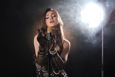 Photo of Beautiful young woman with microphone singing on dark background with smoke