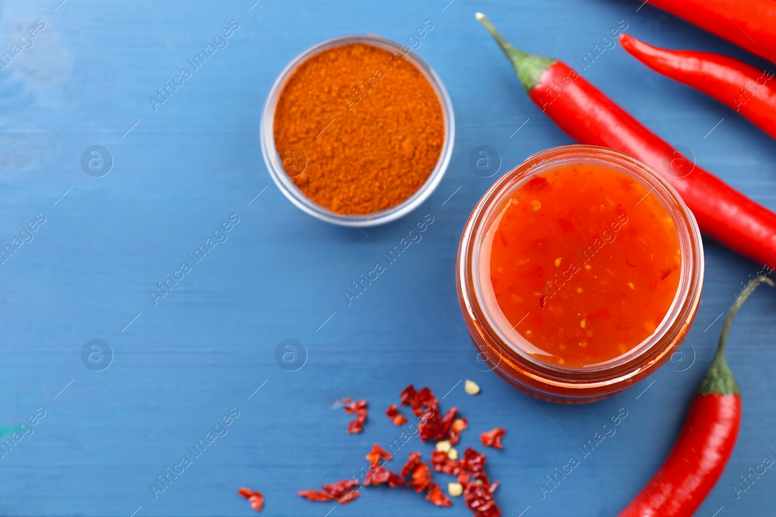 Photo of Spicy chili sauce in jar and ingredients on blue wooden table, flat lay. Space for text