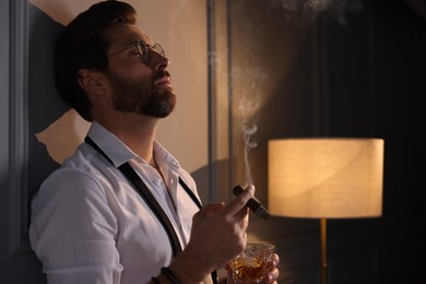 Handsome man with glass of whiskey smoking cigar at home. Space for text