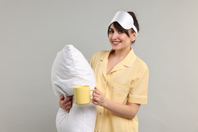 Happy woman in pyjama holding pillow and cup of drink on grey background